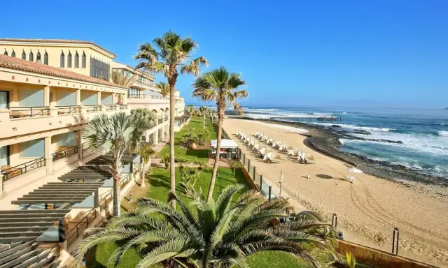 Fuerteventura kiedy wieje, palmy kołyszą się nad piaszczystą plażą i oceanem. Hotel z tarasami i widokiem na fale.