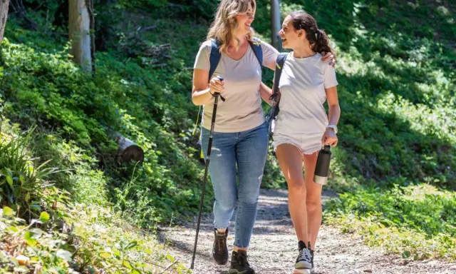 Två glada kvinnor njuter av en promenad i skogen, med en vandringsstav och en vattenflaska.