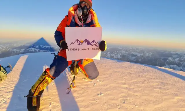 Na szczycie, gdzie jest K2, wspinacz w pomarańczowym kombinezonie trzyma tabliczkę "Seven Summit Treks".