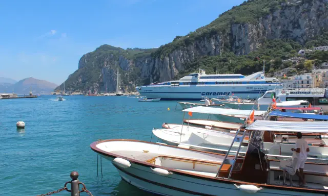 Barche in attesa al porto, con la maestosa isola di Capri sullo sfondo. Un traghetto Caremar attende passeggeri da Napoli.