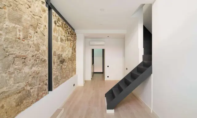 Interior moderno con pared de piedra rústica, escaleras oscuras y suelo de madera clara. Un ejemplo de cómo hacer una casa sostenible combinando lo antiguo y lo nuevo.