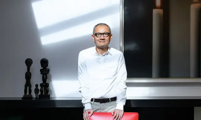 Pierre Chen, a man with glasses, stands in a modern room with striking shadows. He wears a white shirt and tan pants, leaning on a red chair.