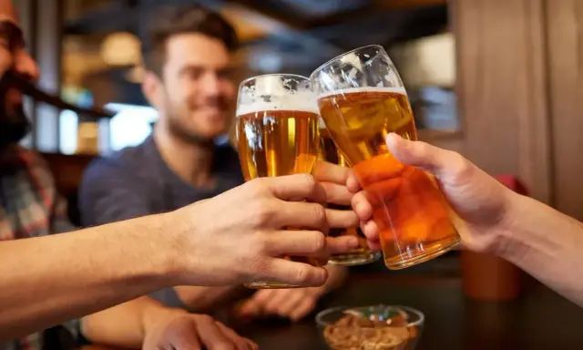Trzech przyjaciół wznosi toast piwem. Widać ich dłonie trzymające szklanki z bursztynowym napojem, a na stole miseczkę z orzeszkami. To idealny moment, by porozmawiać o zawartości alkoholu w piwie.