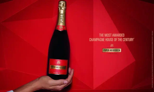 Eine Hand hält eine Flasche Piper-Heidsieck Champagner vor rotem, geometrischem Hintergrund.
