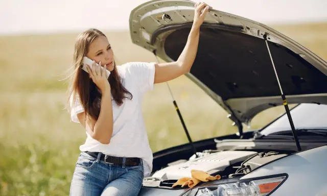 Mujer llama por teléfono con el capó abierto, preocupada por qué pasa si el coche se queda sin aceite.
