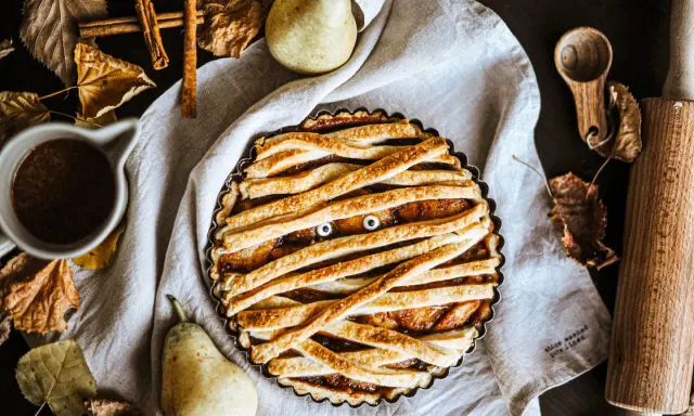 Tarte d'Halloween en forme de momie, avec des yeux globuleux et une garniture aux fruits. Autour, des feuilles d'automne, des poires et des épices.