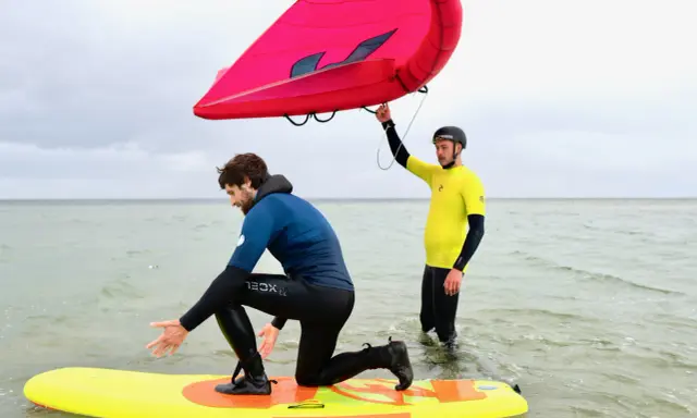Zwei Männer üben Wing Surfen im flachen Wasser. Einer kniet auf einem gelben Board, der andere hält einen roten Wing.