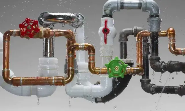 Complejo sistema de varios tipos de tuberías para agua: cobre, PVC, metal y negro, con válvulas rojas y verdes, goteando agua.