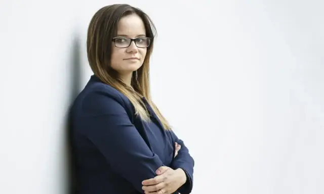 Kobieta w okularach i granatowej marynarce stoi oparta o ścianę, ze skrzyżowanymi rękami.