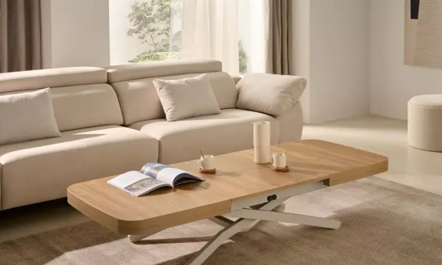 Mesa centro elevable 120x70 de madera clara, con un libro abierto y taza de café, junto a un sofá beige en un salón luminoso.