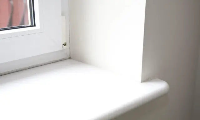 Biały parapet okienny, gotowy do wymiany. Widoczny fragment okna i ściany.