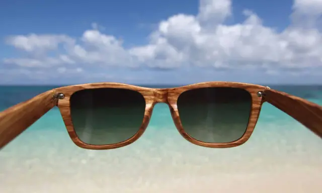 Drewniane okulary przeciwsłoneczne z filtrem uv 3 chronią oczy na plaży. W tle błękitne niebo i turkusowe morze.