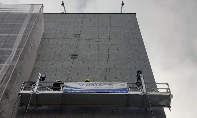 Dos trabajadores en andamios suspendidos realizan labores en la fachada de un edificio.