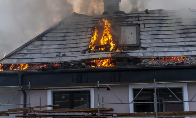 Un **incendie habitation** ravage le toit d'une maison, avec des flammes vives et une épaisse fumée s'échappant des combles.