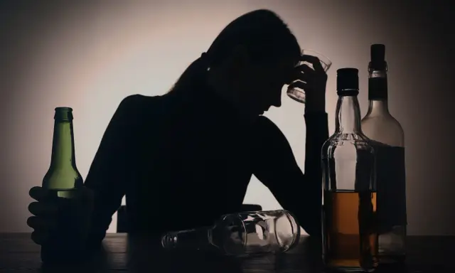 Sylwetka kobiety z kieliszkiem w dłoni, otoczona butelkami. Widoczny problem z przedawkowaniem alkoholu.