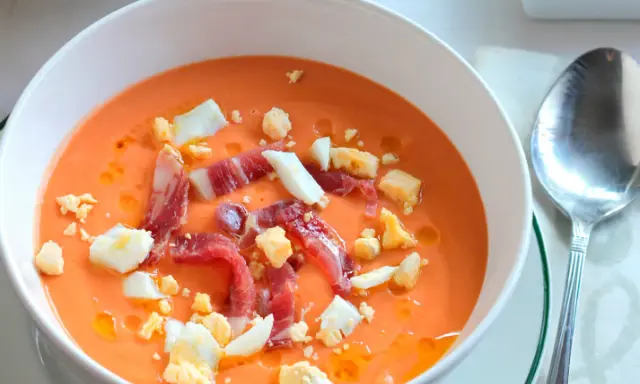 Un cuenco blanco con salmorejo cordobés, coronado con huevo duro picado y taquitos de jamón serrano.