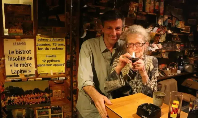 Au café du coin Le Mans, Jeanine, 73 ans, fan de rockeur, allume sa pipe. Un homme souriant se tient à ses côtés.