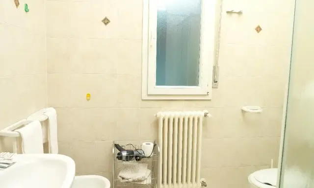 Bagno 79 Rimini: lavabo, bidet, WC, scaldasalviette e finestra. Asciugacapelli e rotolo di carta igienica su carrello.