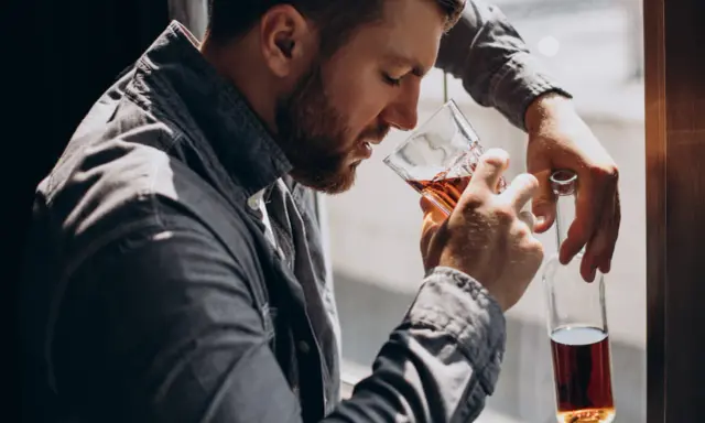 Mężczyzna z brodą, oparty o okno, pije alkohol. W ręku trzyma szklankę z bursztynowym płynem i pustą butelkę. To obraz, jak alkoholik zapije się na śmierć.
