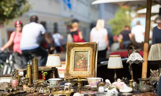 Antikviteter och kuriosa på ett bord, kanske på en loppmarknad i Stockholm. En blomstermålning i guldram, små lampor och porslin.