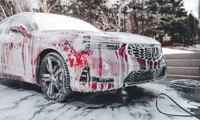 Czerwony samochód pokryty pianą podczas czyszczenia. Widać felgi, reflektory i grill.