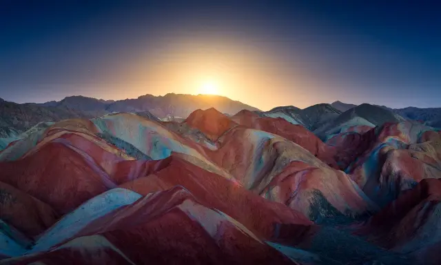 Bunte Schichten aus Rot, Gelb und Blau formen die **Regenbogenberge Chinas** unter einem tiefblauen Himmel mit aufgehender Sonne.