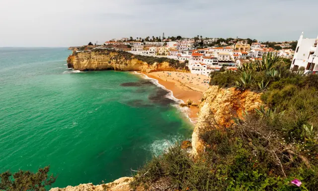 Blick auf die sonnenverwöhnte **Carvoeiro Altstadt** mit ihren weißen Häusern, die sich an die Klippen schmiegen. Der goldene Sandstrand lädt zum Verweilen ein, während das smaragdgrüne Meer sanft an die Küste brandet.