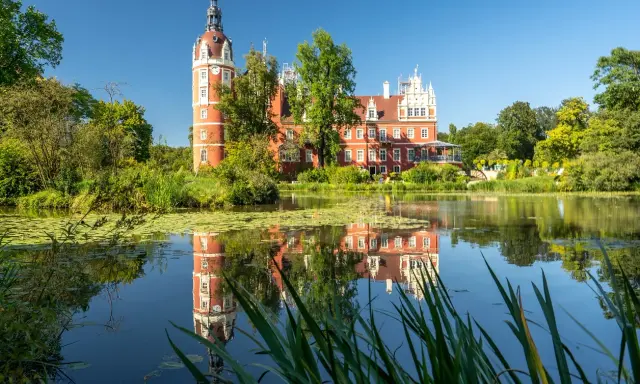 Czerwony zamek z wieżą odbija się w spokojnej tafli jeziora. Malowniczy krajobraz wpisuje się w piękno geoparku Łuk Mużakowa.