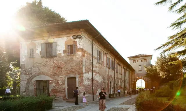 Un sole caldo illumina l'antica **abbazia di Chiaravalle**, con i suoi mattoni a vista e le persiane chiuse. Persone passeggiano nel cortile acciottolato.