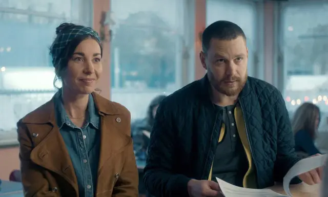 En kvinna med scarf och en man med skägg sitter vid ett bord. De ser ut att vara med i en dansk serie på SVT.