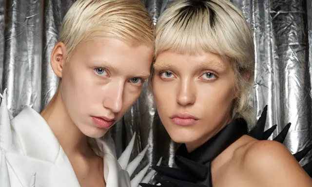 Zwei Models mit Pixie Cut feinem Haar und auffälligen Outfits vor silberner Kulisse.