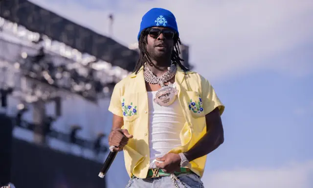 Chief Keef, w niebieskiej czapce i żółtej koszuli z kwiatami, na scenie. Jego wiek nie jest widoczny, ale styl mówi wszystko.