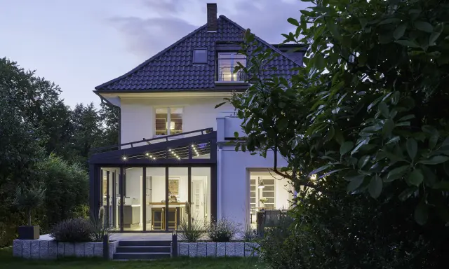 Moderner Anbau Wintergarten mit Glasfront und Beleuchtung erweitert ein Haus. Der Garten ist abends beleuchtet.