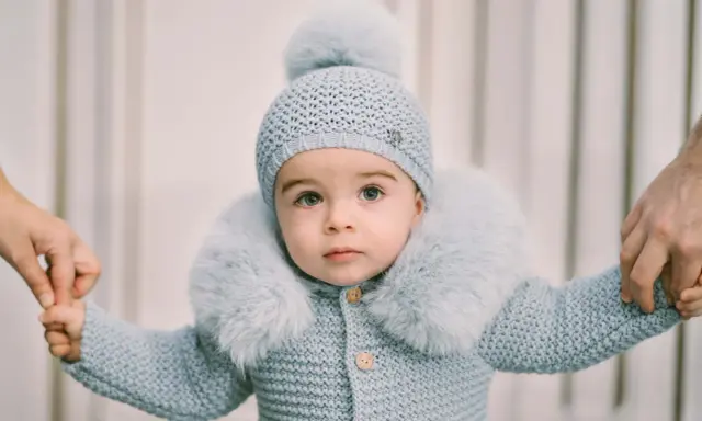 Un tierno bebé con un conjunto de punto azul claro, gorro y abrigo con cuello de pelo. Sus manos son sujetadas por adultos, como si estuviera aprendiendo a caminar. Este patrón de pelele para bebé es adorable.