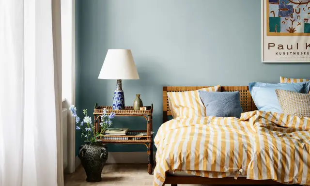 Dormitorio con paredes azul claro y ropa de cama a rayas amarillas y blancas. Los relajantes colores para dormitorios se complementan con una mesita de noche de mimbre y un jarrón con flores.