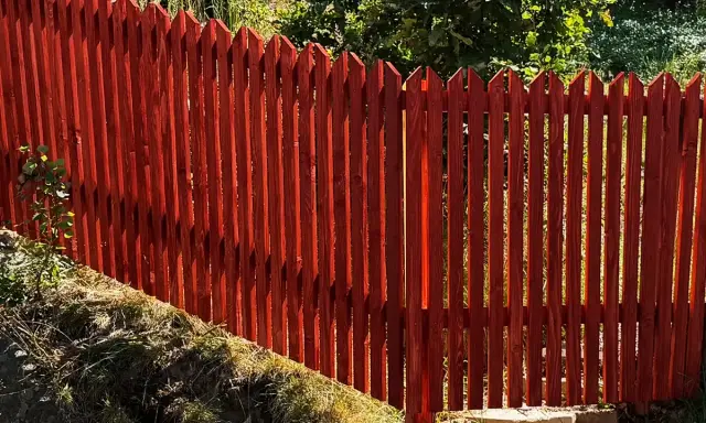 Rött, spetsigt staket av tryckimpregnerat trä. Solen lyser genom plankorna och skapar skuggor.
