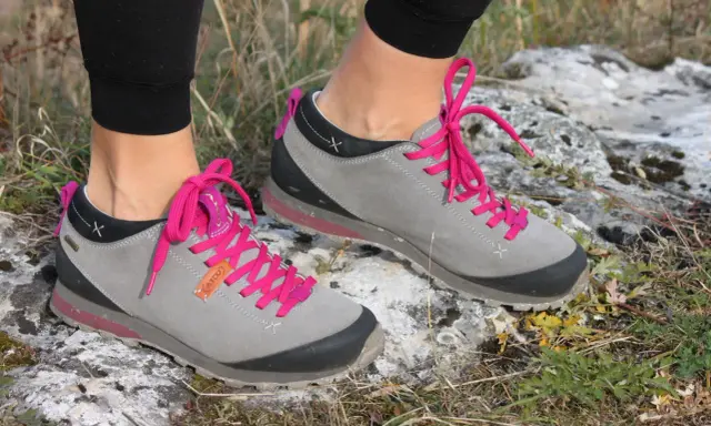 Szare buty trekkingowe Aku Bellamont z jaskrawymi, różowymi sznurówkami na kamienistym, trawiastym podłożu.