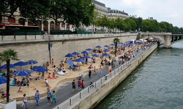 Menschen entspannen sich am **Paris strand** unter blauen Sonnenschirmen. Palmen und Gebäude säumen die Uferpromenade.