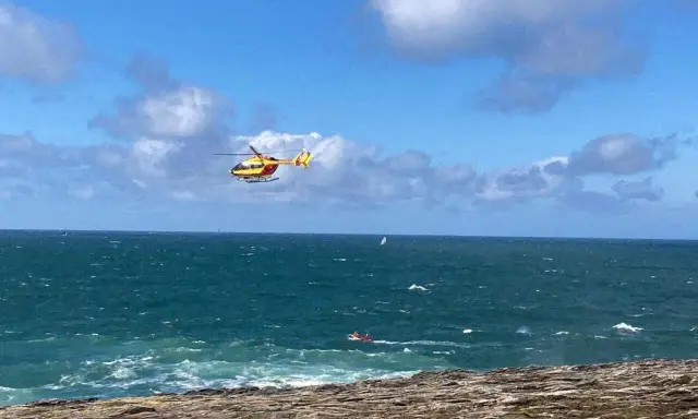 Un hélicoptère jaune et rouge survole une mer agitée, cherchant une personne disparue en mer. Un petit bateau rouge est visible au loin.