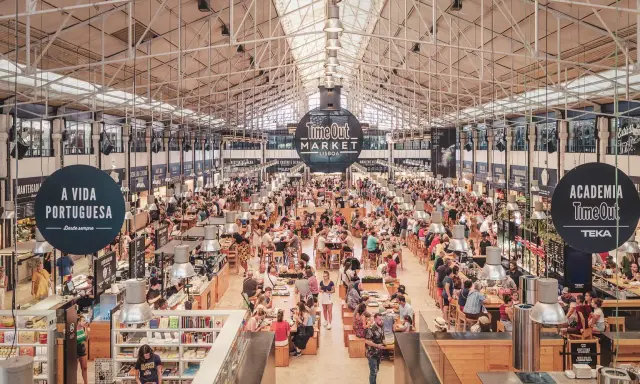 Belebter Time Out Market in Lissabon. Menschen sitzen an langen Tischen, essen und unterhalten sich. Überall gibt es Essensstände.