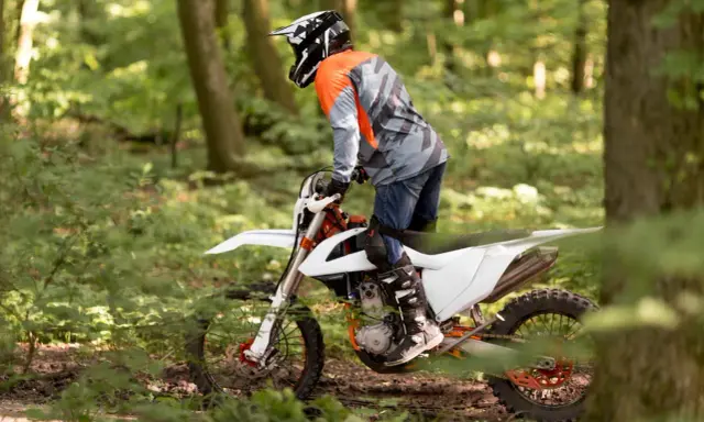 Motocyklista w kasku i pomarańczowej bluzie jedzie na białym crossie przez leśną ścieżkę.