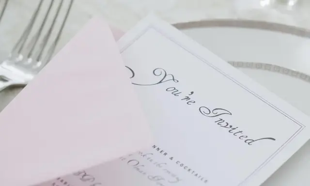 Eleganckie zaproszenie z osobą towarzyszącą, "You're Invited", na różowej kopercie, obok sztućców i talerza.