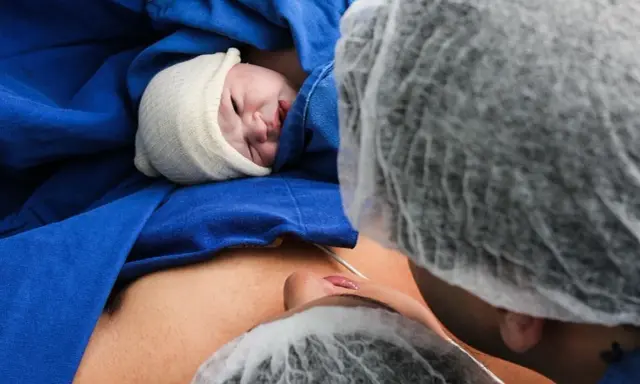 Noworodek w czepku, otulony niebieskim materiałem, leży na brzuchu mamy. Widoczna twarz dziecka i fragment twarzy mamy. To piękny moment porodu lotosowego.