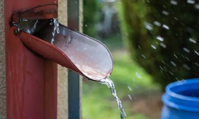 Agua de lluvia cayendo de una canaleta roja hacia un barril azul, una forma práctica de saber como recoger agua de lluvia en un terreno.