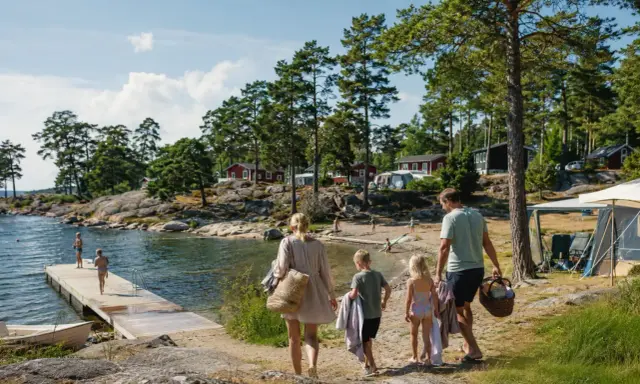 Familj på väg mot vattnet för roliga aktiviteter barn. Soligt, skärgårdsmiljö med röda stugor och camping.