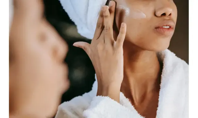 Une femme applique une crème pour l'hydratation du visage, enveloppée dans une serviette et un peignoir douillet.