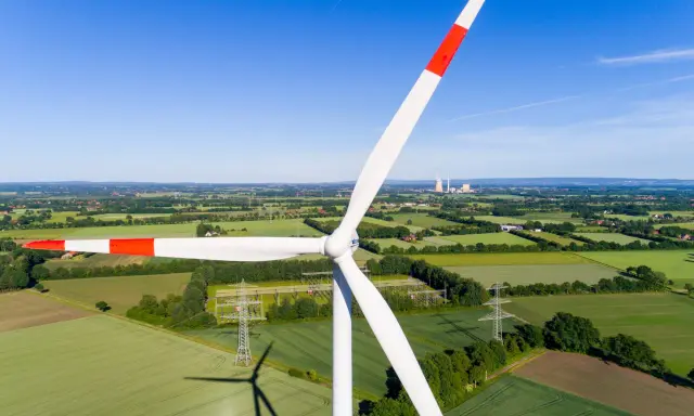 Windkraft Energie: Ein riesiger Windpark dominiert die Landschaft, umgeben von grünen Feldern und Bäumen. Im Hintergrund sind Kühltürme zu sehen.