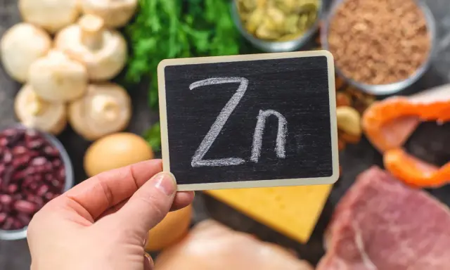 Tabliczka z napisem "Zn" na tle produktów bogatych w cynk: mięso, ryby, fasola, grzyby, jajka, ser, nasiona.