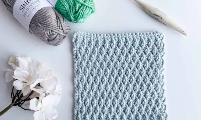 Muestra de tejido a crochet con el patrón nudo de salomon en color azul claro, junto a ovillos de lana gris y verde, una flor blanca y un ganchillo.