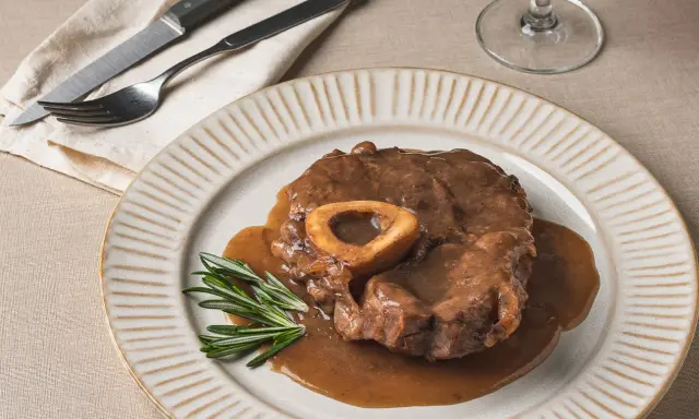 Un delicioso osso buco, tierno y jugoso, bañado en su salsa, servido en un plato con romero fresco.